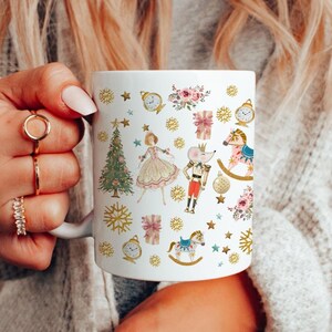 May include: White ceramic mug with a Christmas-themed design featuring a ballerina, a nutcracker, a rocking horse, a Christmas tree, and other festive elements. The design is in shades of pink, gold, and blue.
