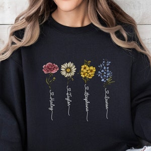 Pu&ograve; includere: Felpa nera con un design floreale e i nomi "Hailey", "Taylor", "Stephanie" e "Lauren" scritti verticalmente sotto i fiori.