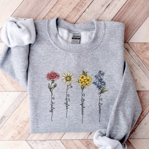Pu&ograve; includere: Felpa grigia con un design floreale e i nomi "Hailey", "Taylor", "Stephanie" e "Lauren" scritti sotto i fiori.
