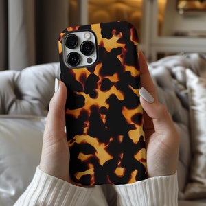 May include: A close-up of a phone case with a tortoiseshell pattern. The case is brown and black with a yellow-orange swirl. The phone case is being held by a person's hands.