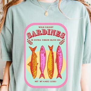 以下が含まれることがあります： レトロスタイルのイワシのラベルデザインが特徴のライトブルーのTシャツ。ピンクとグリーンのフレームに入ったラベルには、「Sardines」の文字と4匹のカラフルな魚のイラストが描かれています。「Wild-Caught」と「In Extra Virgin Olive Oil」の文字も表示されています。