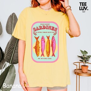 以下が含まれることがあります： レトロなイワシ缶のデザインが特徴のイエローTシャツ。「WILD-CAUGHT SARDINES IN EXTRA VIRGIN OLIVE OIL」の文字と4匹の様式化された魚が描かれています。正味重量は125gです。ブランドロゴ「TEE LUV」が右上隅にあります。