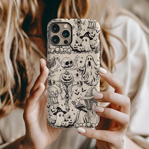 May include: Beige phone case with a black and white Halloween-themed pattern. The design includes ghosts, pumpkins, witches, skulls, and bats. The case is held by a person.