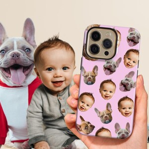 May include: A pink phone case featuring a pattern of baby faces and French bulldog faces. The case is held in a hand, with a baby and a French bulldog in the background. The dog is wearing a red and white outfit.