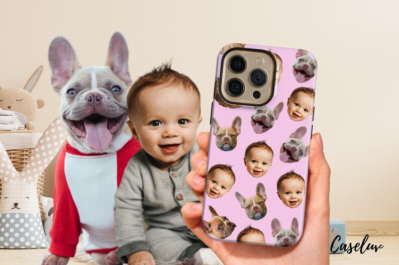 May include: A pink phone case featuring a pattern of baby faces and French bulldog faces. The case is held in a hand, with a baby and a French bulldog in the background. The dog is wearing a red and white outfit.