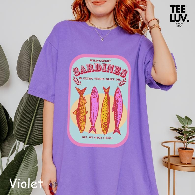 以下が含まれることがあります： 紫色のTシャツ。エキストラバージンオリーブオイル漬けのイワシのグラフィックが特徴です。デザインには、水色とピンクの長方形のフレームの中に、4匹のカラフルな魚のイラストが含まれています。「Wild-Caught Sardines」の文字が魚の上に、「Net Wt 125g」が下にあります。