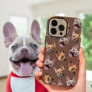 May include: A brown phone case featuring a pattern of French bulldog faces. The phone case is held in front of a smiling French bulldog with a red and white shirt. The phone case has a gold-colored camera ring.
