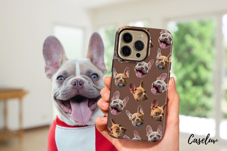 May include: A brown phone case featuring a pattern of French bulldog faces. The phone case is held in front of a smiling French bulldog with a red and white shirt. The phone case has a gold-colored camera ring.