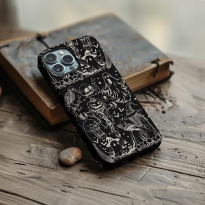 May include: Black phone case with a Halloween-themed design. The case features a detailed white illustration of ghosts, pumpkins, bats, and a witch. The case is resting on a wooden surface next to a book.