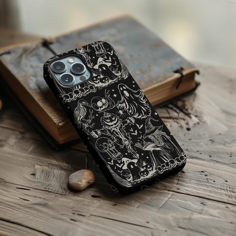 May include: Black phone case with a Halloween-themed design. The case features a detailed white illustration of ghosts, pumpkins, bats, and a witch. The case is resting on a wooden surface next to a book.