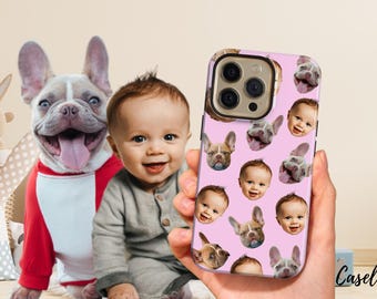 May include: A pink phone case featuring a pattern of baby faces and French bulldog faces. The case is held in a hand, with a baby and a French bulldog in the background. The dog is wearing a red and white outfit.
