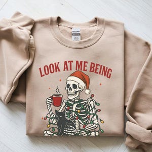 May include: Beige sweatshirt with the text "LOOK AT ME BEING". The graphic features a skeleton wearing a Santa hat, holding a mug, and a black cat. The skeleton is decorated with Christmas lights.