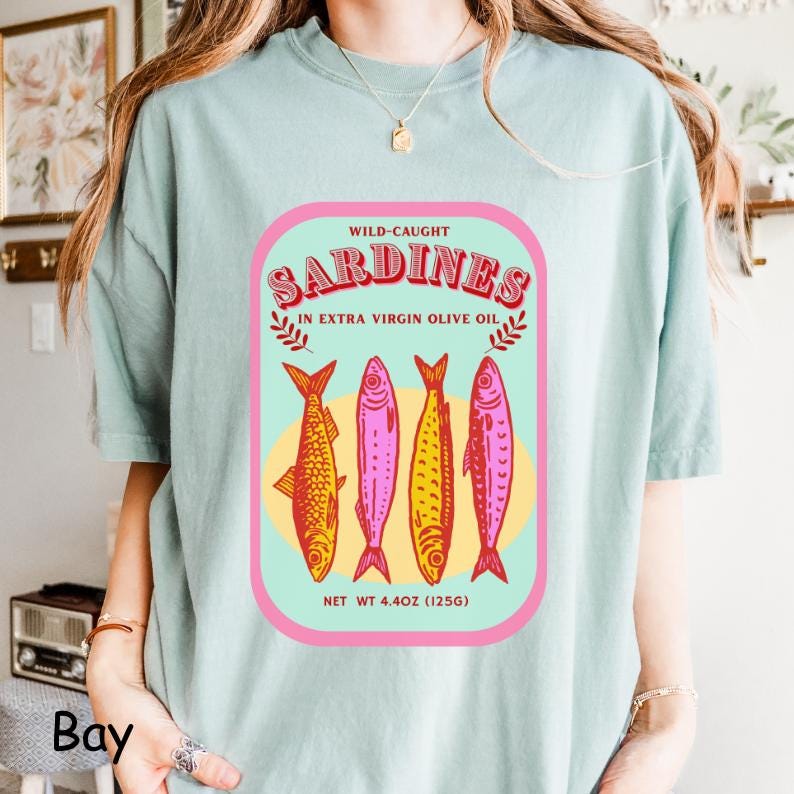 以下が含まれることがあります： レトロスタイルのイワシのラベルデザインが特徴のライトブルーのTシャツ。ピンクとグリーンのフレームに入ったラベルには、「Sardines」の文字と4匹のカラフルな魚のイラストが描かれています。「Wild-Caught」と「In Extra Virgin Olive Oil」の文字も表示されています。