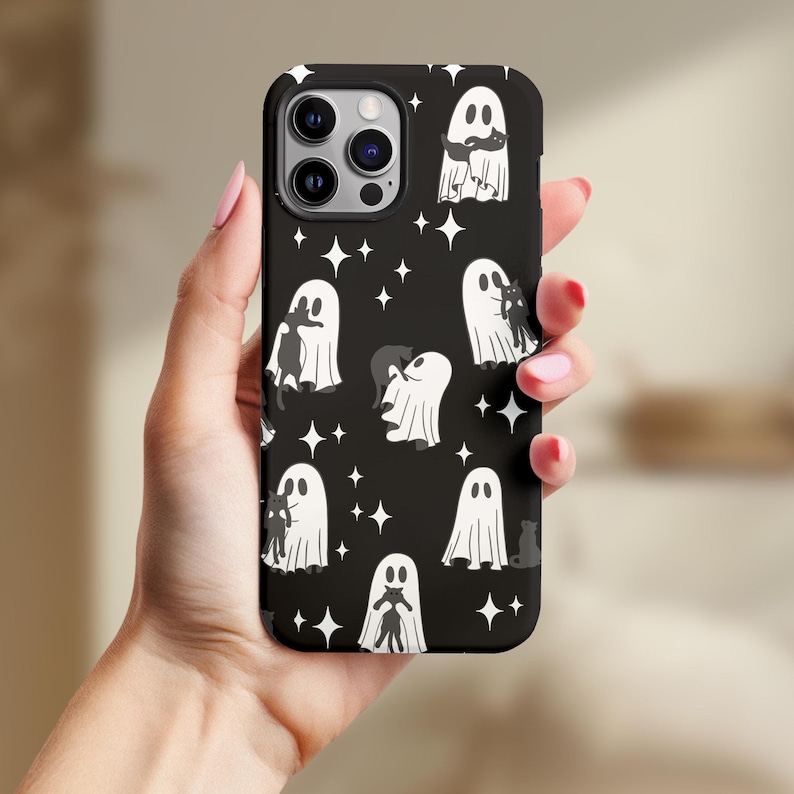 May include: Black phone case with a repeating pattern of white ghosts and black cats, interspersed with white star shapes. The case has a glossy finish and is held in a hand.