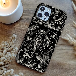 May include: Black phone case with a Halloween-themed design. The case features white illustrations of ghosts, pumpkins, bats, skulls, and a witch. The case is on a wooden surface with a lit candle and decorative plants.