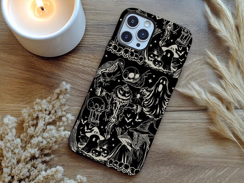 May include: Black phone case with a Halloween-themed design. The case features white illustrations of ghosts, pumpkins, bats, skulls, and a witch. The case is on a wooden surface with a lit candle and decorative plants.