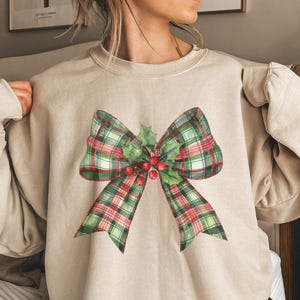 May include: A beige sweatshirt featuring a watercolor illustration of a Christmas bow. The bow has a green and red plaid pattern, with holly leaves and red berries in the center. The sweatshirt is a neutral color.