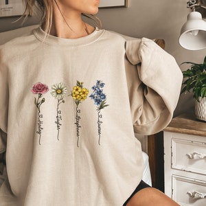 Pu&ograve; includere: Una felpa beige con un design floreale e quattro nomi scritti sotto ogni fiore. I nomi sono "Hailey", "Taylor", "Stephanie" e "Lauren".