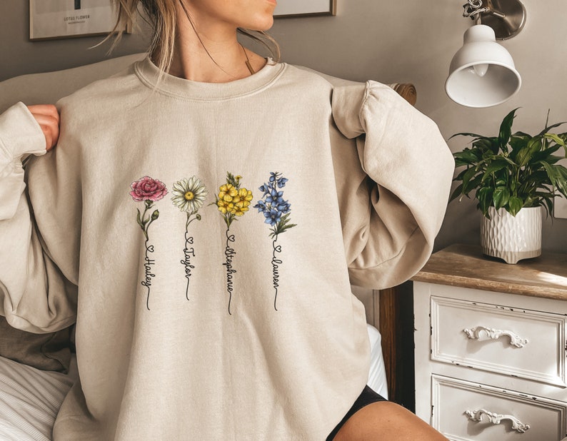 Pu&ograve; includere: Una felpa beige con un design floreale e quattro nomi scritti sotto ogni fiore. I nomi sono "Hailey", "Taylor", "Stephanie" e "Lauren".