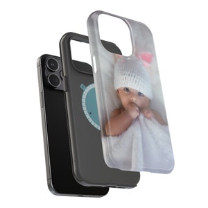 Peut inclure: Un smartphone noir avec une coque de protection repr&eacute;sentant un b&eacute;b&eacute; portant un bonnet blanc tricot&eacute; avec des accents roses. La coque est transparente et le b&eacute;b&eacute; est envelopp&eacute; dans une couverture blanche.