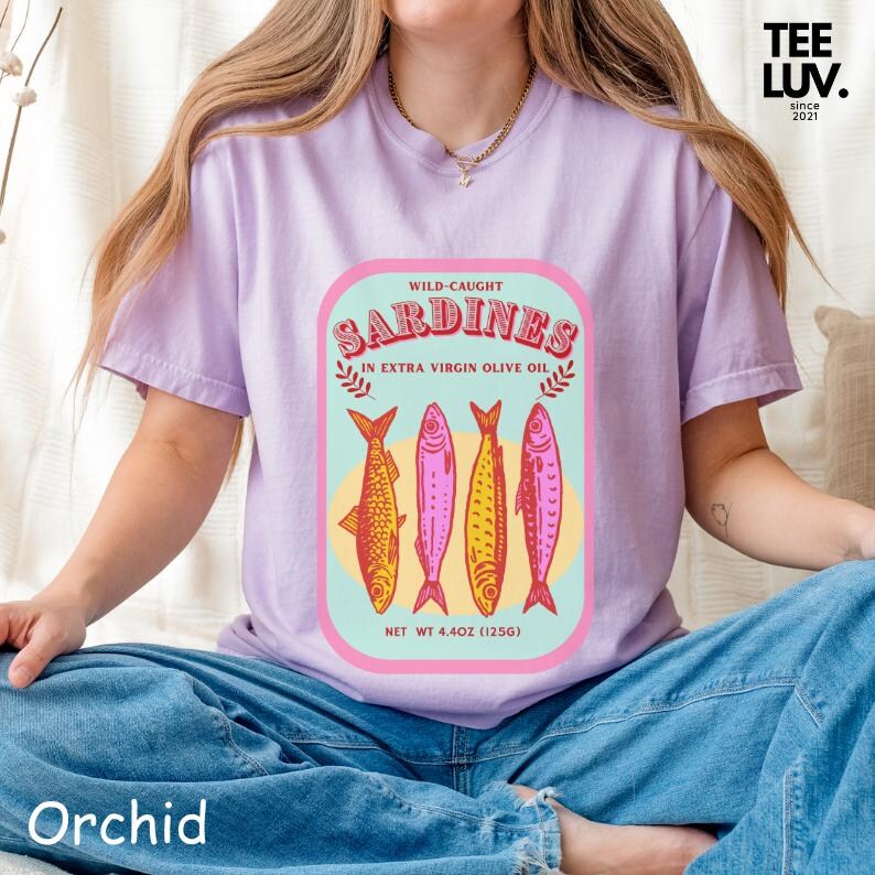 以下が含まれることがあります： 紫色のTシャツ。イワシの缶詰のデザインが特徴です。「Sardines」の文字はレトロなスタイルで、4匹のカラフルな魚のイラストが描かれています。「Wild-Caught」と「In Extra Virgin Olive Oil」の文字も含まれています。Tシャツには「TEE LUV」と「since 2021」の文字があります。