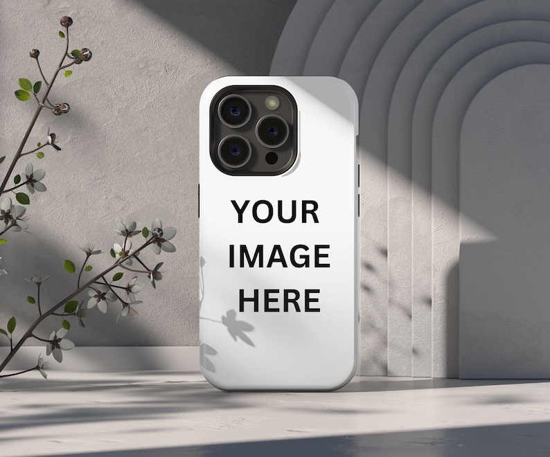 Peut inclure: Coque de t&eacute;l&eacute;phone blanche avec module cam&eacute;ra noir et le texte "YOUR IMAGE HERE". L'&eacute;tui est pr&eacute;sent&eacute; dans un studio avec un fond neutre et une branche de fleurs.