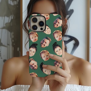 May include: Green phone case featuring a repeating pattern of various faces. The case has a camera cutout and is held up in front of a person. The faces include adults and babies, with a variety of expressions.