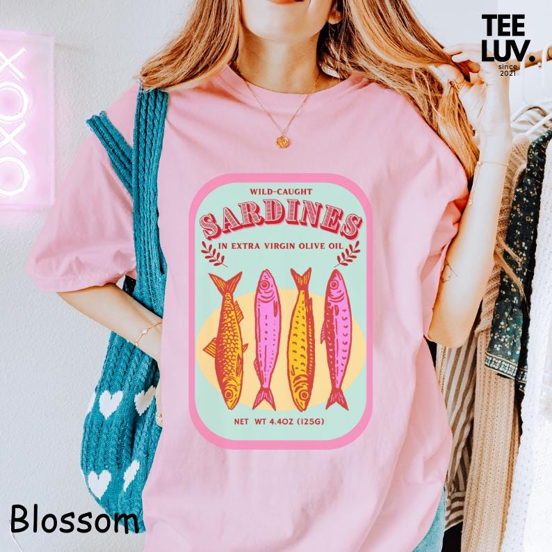 以下が含まれることがあります： ピンク色のTシャツ。イワシの缶詰ラベルのデザインで、「WILD-CAUGHT SARDINES IN EXTRA VIRGIN OLIVE OIL」と書かれ、4匹の魚のイラストが描かれています。Tシャツには「Blossom」と「TEE LUV since 2021」の文字も入っています。