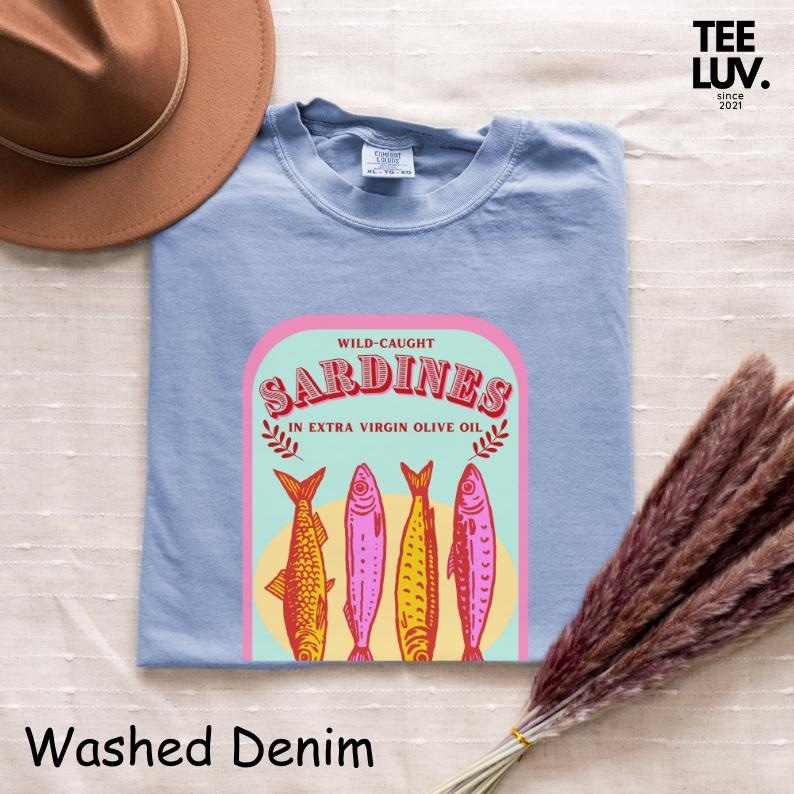 以下が含まれることがあります： ウォッシュデニムのライトブルーTシャツ。エキストラバージンオリーブオイル漬けのイワシのグラフィックが特徴です。グラフィックには、4匹のカラフルな魚のイラストが描かれています。シャツには「WILD-CAUGHT SARDINES」と「TEE LUV. since 2021」の文字が入っています。