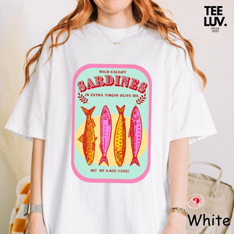 以下が含まれることがあります： カラフルなイワシのデザインが施された白いTシャツ。「WILD-CAUGHT SARDINES IN EXTRA VIRGIN OLIVE OIL」と「NET WT 125g」の文字が含まれています。さまざまな色の4匹のイワシが描かれています。