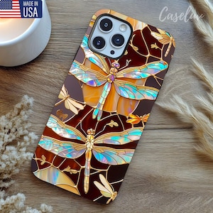 May include: A phone case with a stained-glass dragonfly design. The case features a brown background with gold, blue, and orange dragonfly illustrations. The case has a glossy finish and is made in the USA.