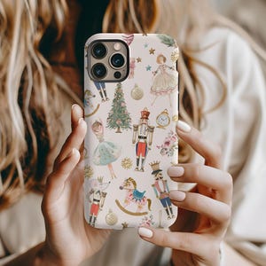 May include: A white phone case with a Christmas Nutcracker theme. The case features a repeating pattern of Nutcracker soldiers, ballerinas, Christmas trees, and other festive elements. The case is designed for an iPhone.