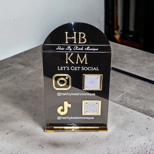 May include: Black acrylic social media sign with gold lettering and accents. The sign features the text "HB Hair By Keish Monique KM LET'S GET SOCIAL" along with Instagram, TikTok, and QR code icons. The Instagram and TikTok handles are also displayed.