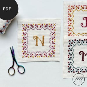 Peut inclure: Broderie au point de croix avec la lettre "N" dorée, encadrée d'une bordure décorative violette, dorée et rouge. Le motif est sur un tissu blanc, avec de petits ciseaux colorés. D'autres échantillons de point de croix sont visibles.