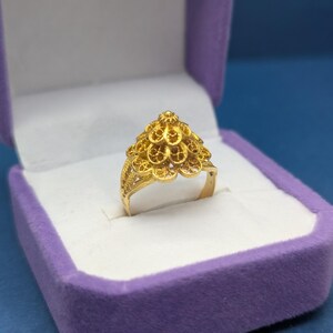 May include: A gold ring with a delicate, intricate design. The ring is in a purple velvet ring box.