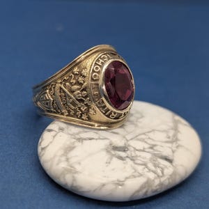 May include: A gold-toned class ring with a large, oval, deep red gemstone. The ring features intricate side detailing and the inscription "RITA SCHOOL". The ring is displayed on a white and gray marbled stone against a blue background.