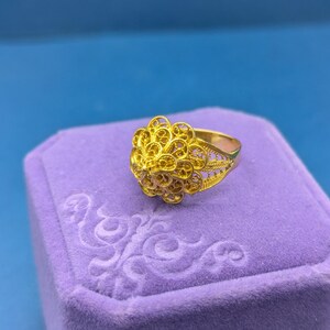 May include: A gold filigree ring with a delicate floral design. The ring is displayed in a purple velvet ring box.