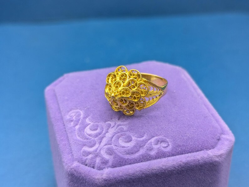 May include: A gold filigree ring with a delicate floral design. The ring is displayed in a purple velvet ring box.