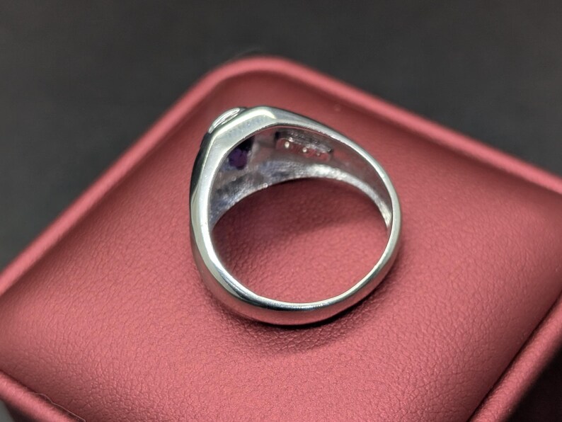 May include: A silver ring with a rounded band and a rectangular face. The face of the ring features a small, round, purple gemstone and a smaller, clear gemstone. The ring is set on a red surface.