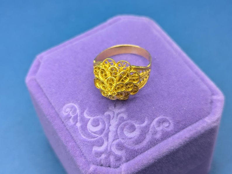 May include: A gold filigree ring with a delicate, intricate design. The ring is displayed in a purple velvet ring box.