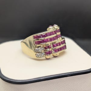 May include: A gold ring with a rectangular design, featuring rows of square-cut rubies and small diamonds. The ring has a vintage style and is set on a white surface with a black background.