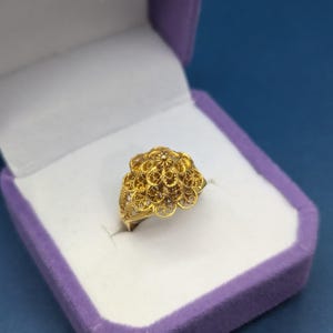 May include: A gold filigree ring with a flower-shaped design. The ring is displayed in a purple velvet ring box.