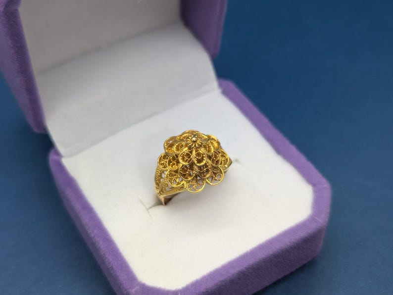 May include: A gold filigree ring with a flower-shaped design. The ring is displayed in a purple velvet ring box.