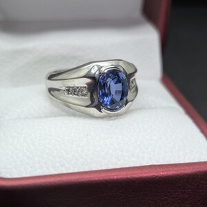 May include: A silver ring with a large oval blue gemstone at the center. The ring has a modern design with small accent stones on each side of the main stone. The ring is displayed in a red jewelry box.