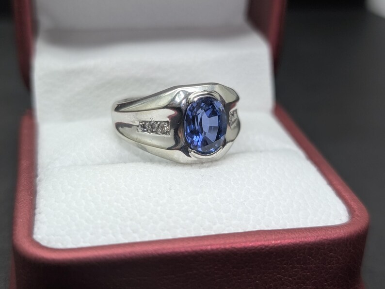 May include: A silver ring with a large oval blue gemstone at the center. The ring has a modern design with small accent stones on each side of the main stone. The ring is displayed in a red jewelry box.