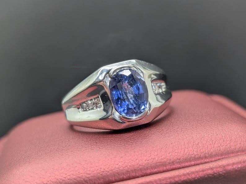 May include: A silver ring with a large, oval, blue gemstone as the centerpiece. Small, clear stones are set on either side of the main stone. The ring is displayed on a pink surface.