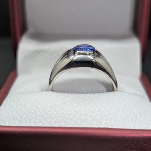 May include: A silver ring with a blue gemstone, presented in a red and white jewelry box. The ring features a polished band and a rectangular setting for the oval-shaped blue stone. The box has a textured interior.