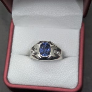 May include: A silver ring with a large oval blue gemstone at the center, flanked by smaller clear stones. The ring is displayed in a red and white jewelry box, showcasing its design and craftsmanship.