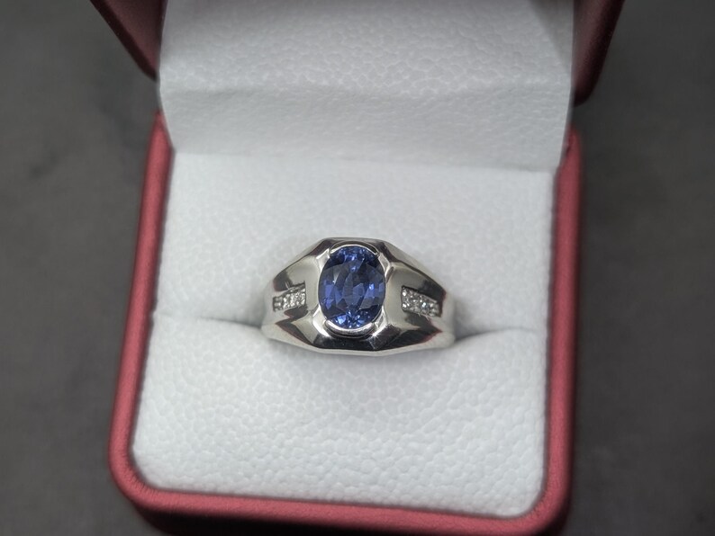 May include: A silver ring with a large oval blue gemstone at the center, flanked by smaller clear stones. The ring is displayed in a red and white jewelry box, showcasing its design and craftsmanship.