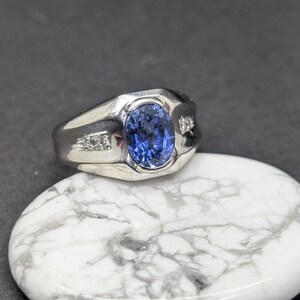 May include: A silver ring with a large oval blue gemstone at the center. Small clear stones are set on either side of the main stone. The ring is displayed on a white and gray marbled surface.